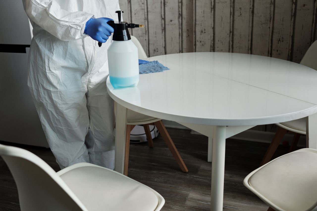 An individual in a protective suit is disinfecting a white table indoors using a sprayer.